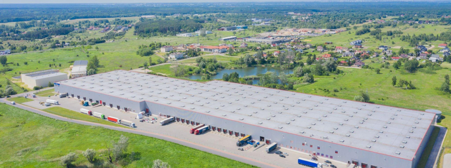 Aerial view of a long warehouse with trucks at loading bays and green surroundings.