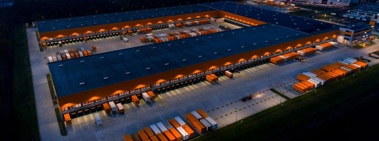 Aerial night view of illuminated warehouse loading bays and parked trucks.