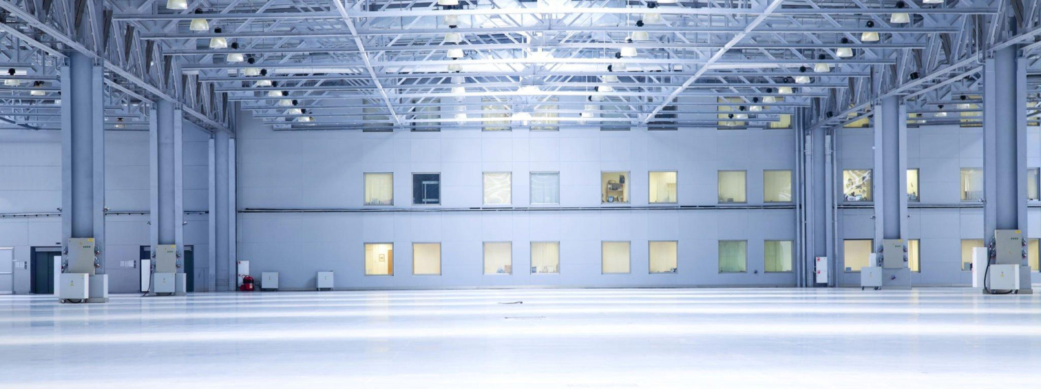 Empty industrial hall with steel roof trusses, overhead lighting and high windows.